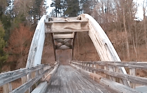 A wooden arch bridge spans a quiet path, surrounded by trees and a serene landscape.