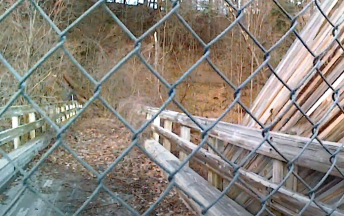 A wooden structure leaning against a hillside, viewed through a chain-link fence, surrounded by dry leaves and trees.