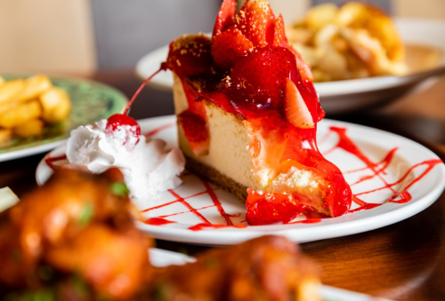 A slice of strawberry cheesecake topped with strawberries, cherry, and whipped cream on a plate, with food in the background.