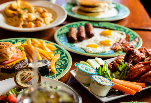 A table spread with a burger, fries, pancakes, eggs, sausage, wings, salad, and dipping sauces.