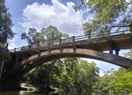 A concrete arch bridge spans over a river, surrounded by lush green trees and a bright blue sky with clouds.