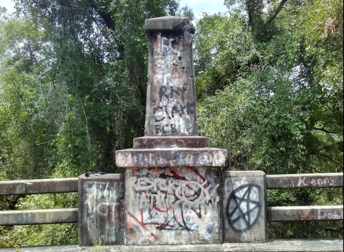 Graffiti-covered stone pillar surrounded by dense greenery, with various tags and symbols visible on the surface.