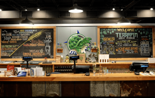 A bar counter with chalkboard menus, a turtle logo, and various beer taps in a cozy brewery setting.