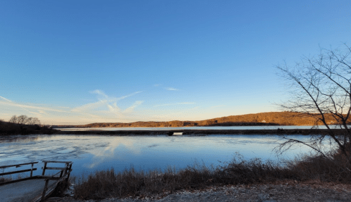 A serene river landscape under a clear blue sky, with trees and distant hills reflecting on the water's surface.