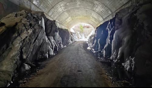 A dimly lit tunnel with rocky walls and a dirt path leading to a bright exit at the end.
