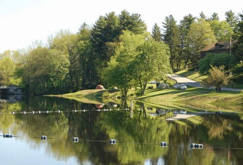 A serene lake scene with trees, tents, and a house reflected in the water under clear skies.
