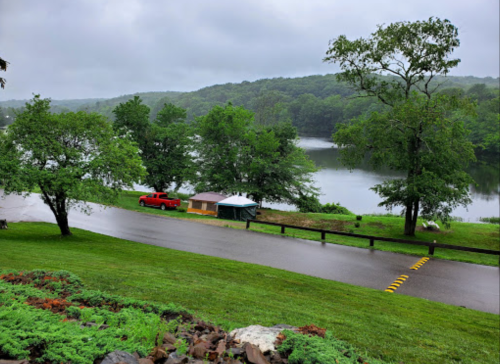 A serene lakeside view with green trees, a red truck, and a small shelter under a cloudy sky.