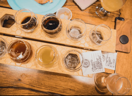 A wooden flight board with eight small glasses of different craft beers on a table.