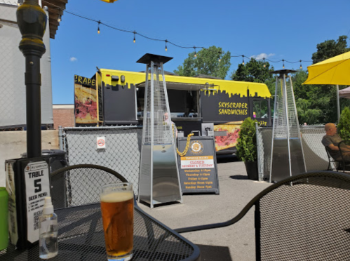 A food truck with a yellow awning serves sandwiches, with outdoor seating and a beer in the foreground.