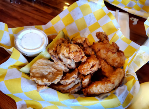 A basket of fried chicken pieces with a side of dipping sauce and pickles, lined with yellow checkered paper.