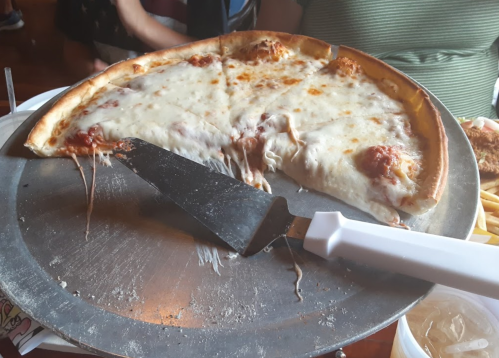 A large cheese pizza on a metal tray, with a slice pulled away, showing melted cheese and a pizza cutter beside it.