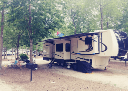 A parked RV in a wooded campsite, with a picnic area and a flag flying above it.