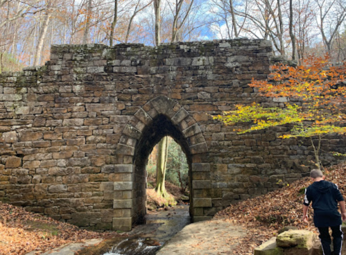 A stone archway bridge surrounded by autumn trees and a person walking nearby on a leaf-covered path.