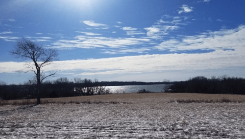 A serene landscape featuring a lake, bare trees, and a clear blue sky with scattered clouds. Snow covers the ground.