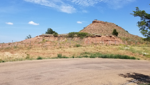 A dry, rocky hillside under a clear blue sky, with sparse vegetation and a paved road in the foreground.