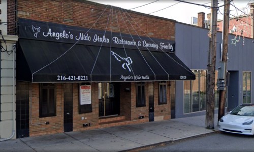 Exterior of Angelo's Nido Italia restaurant, featuring a black awning and brick facade, with a white car parked nearby.