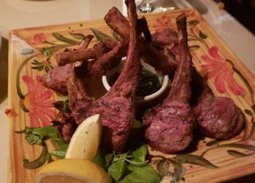 A colorful plate featuring grilled lamb chops, garnished with greens and lemon wedges, served with a small bowl of sauce.