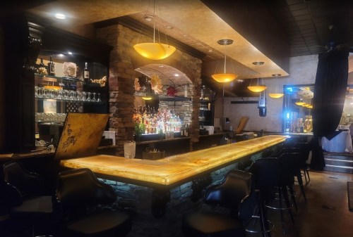 A stylish bar with a glowing countertop, stone accents, and shelves filled with bottles, illuminated by warm lighting.