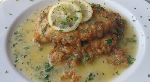 Fried fish fillets topped with lemon slices and herbs, served in a flavorful sauce on a white plate.