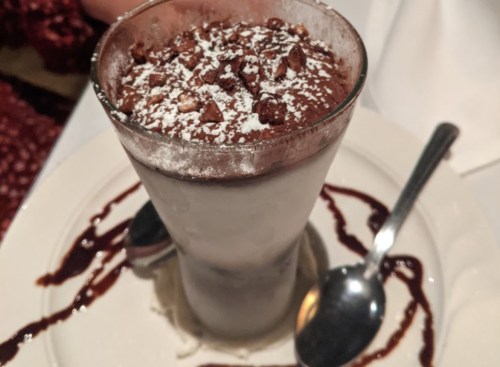 A tall dessert glass filled with layered chocolate mousse, topped with crushed nuts and powdered sugar, on a plate with chocolate drizzle.