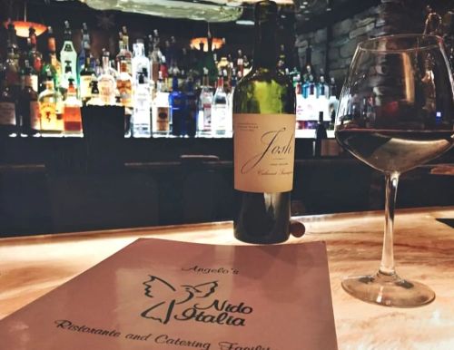 A wine bottle and glass sit on a bar, with a menu for Angelo's Nido Italia and a blurred backdrop of liquor bottles.