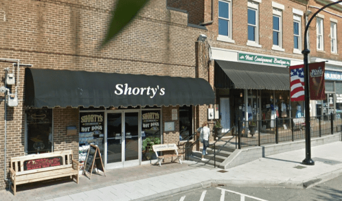 A street view of Shorty's Hot Dogs with a black awning, next to a boutique and an American flag in the background.