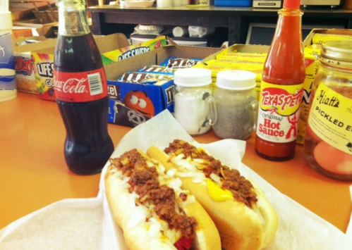 Two chili cheese hot dogs with onions, a bottle of Coca-Cola, and condiments on a table with snacks in the background.