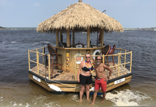 A couple stands on the beach in front of a tiki-style floating bar with a thatched roof.