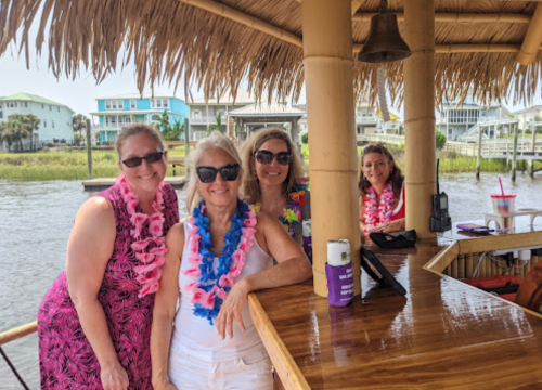 Four women in tropical attire pose at a bar under a thatched roof, with a waterfront view in the background.