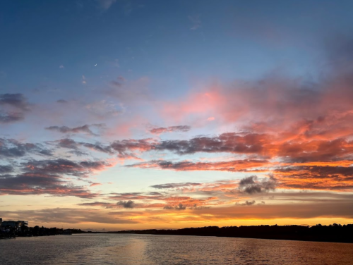 A vibrant sunset over a calm river, with colorful clouds reflecting in the water and a crescent moon in the sky.