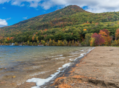 Scenic view of a lake with a sandy shore, surrounded by colorful autumn foliage and a mountain in the background.