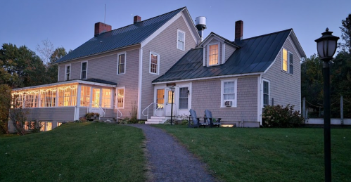 A charming two-story house at dusk, with warm lights glowing from the windows and a grassy path leading to it.