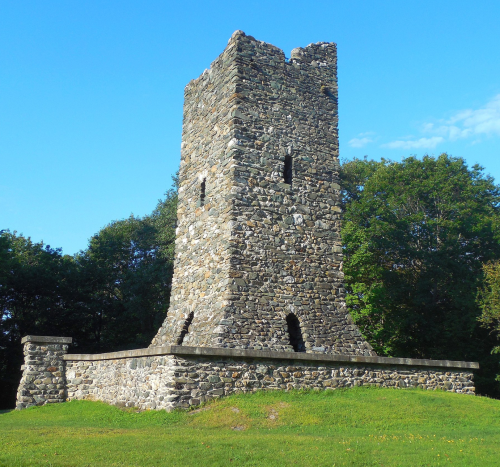 A tall, stone tower stands on a grassy hill, surrounded by trees under a clear blue sky.