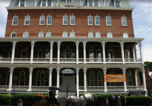 Historic brick building with a large porch, labeled "Museum," and a horse-drawn carriage in front.