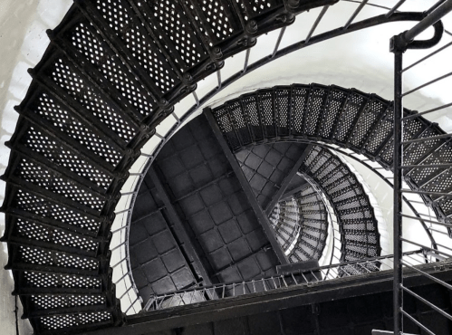 A spiral staircase with black metal steps and railings, viewed from above, creating a dynamic geometric pattern.