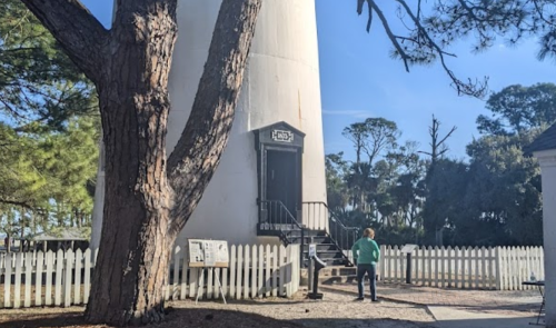 A person stands near a tall lighthouse surrounded by trees and a white picket fence on a sunny day.