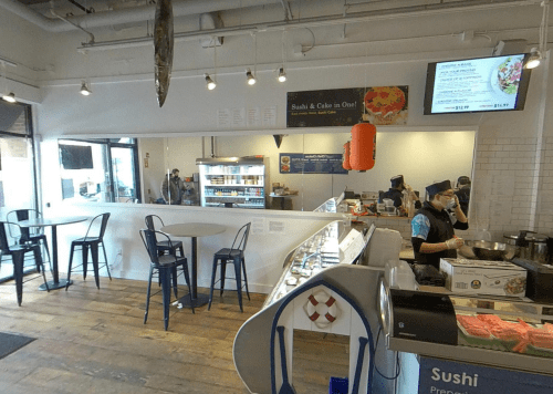 A bright sushi shop with a counter, display cases, and seating, featuring staff preparing food and a menu on the wall.