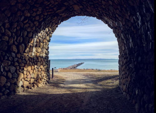 View through a stone archway to a calm lake and a distant pier under a cloudy sky.