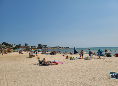 A sunny beach scene with people relaxing on sand, sunbathing, and swimming in the calm water. Beach umbrellas are scattered around.