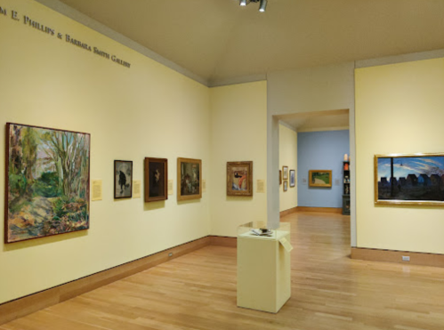 Art gallery interior featuring various framed paintings on yellow walls and wooden flooring.