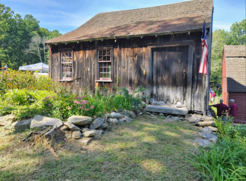 A rustic wooden building with a stone path, surrounded by greenery and flowers, under a clear blue sky.