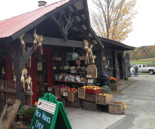 A rustic farm stand with produce, corn decorations, and a sign for new hours, set against a scenic autumn backdrop.
