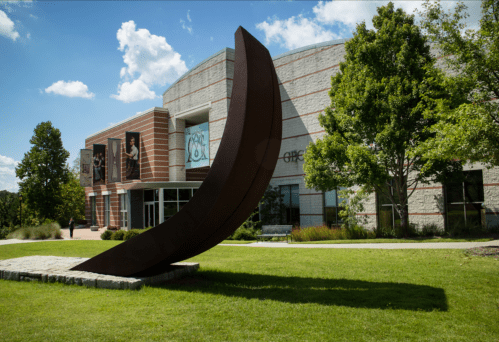 A large curved metal sculpture stands on grass in front of a modern building with trees and blue sky in the background.