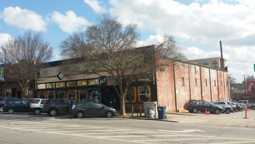 A brick building with large windows, surrounded by parked cars and trees, under a partly cloudy sky.