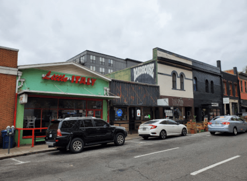 A street view featuring various shops, including "Little Italy," with parked cars along the road under a cloudy sky.