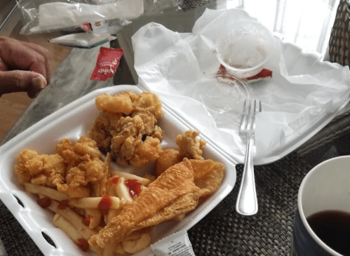 A takeout container with fried chicken, fish, fries, and a small cup, alongside a drink and some condiments on a table.