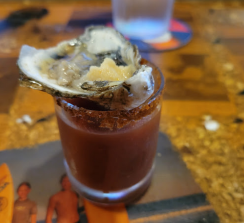 A shot glass filled with a spicy drink topped with an oyster and a salted rim, placed on a wooden table.