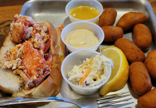 A lobster roll on a bun with sides of coleslaw, dipping sauces, and golden corn fritters on a metal tray.