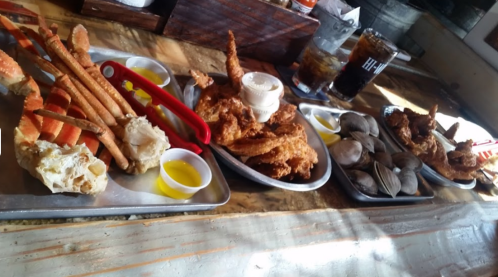 A table with trays of seafood, including crab legs, fried chicken, and clams, accompanied by dipping sauces.
