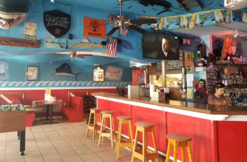 Colorful beach-themed bar interior with a TV, stools, and decor featuring sharks and various brands.